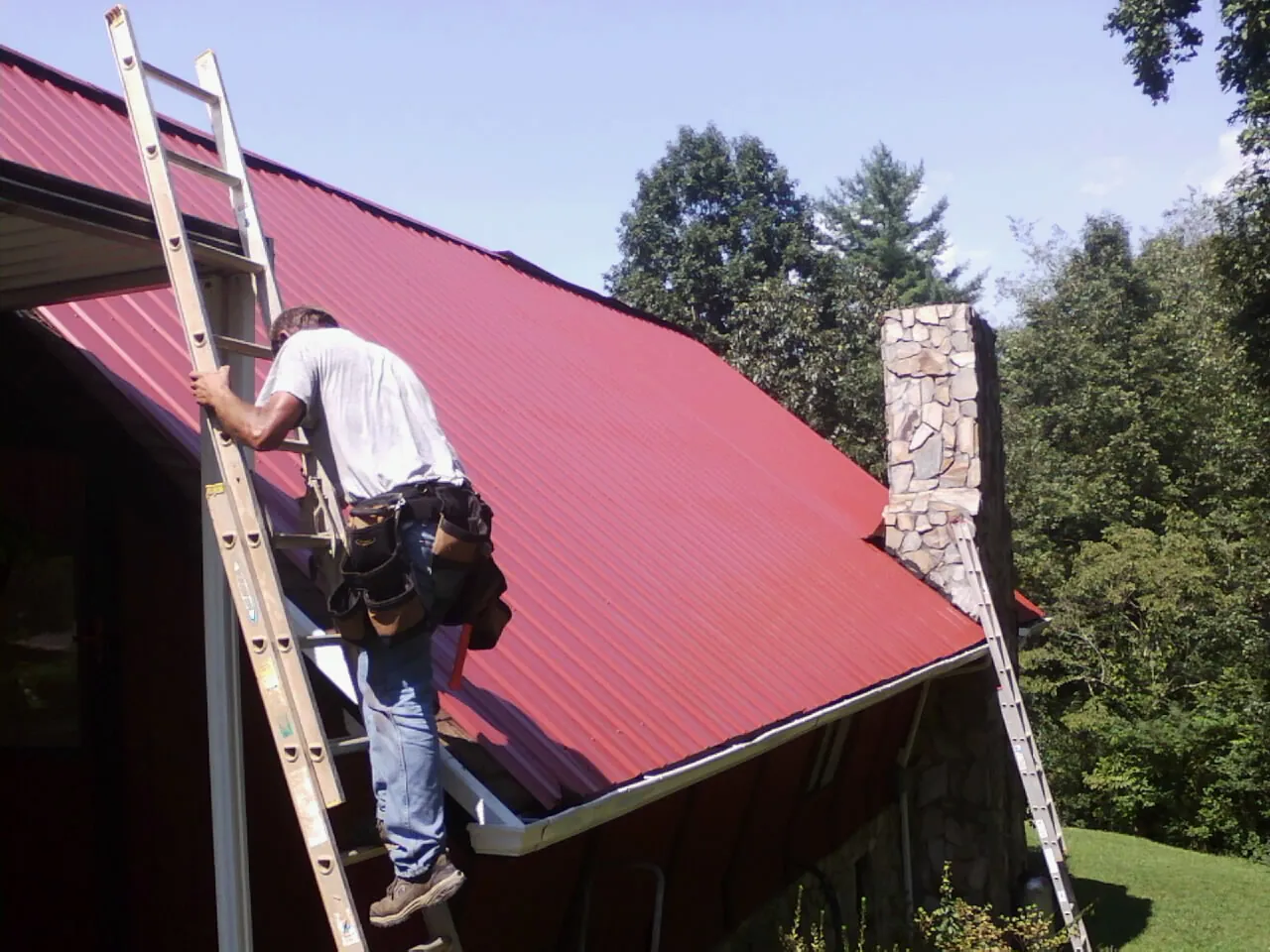 Roofer on a ladder beside a red metal roof and stone chimney — Soffit & Fascia Repair by Roofing Brothers in Hurleyville